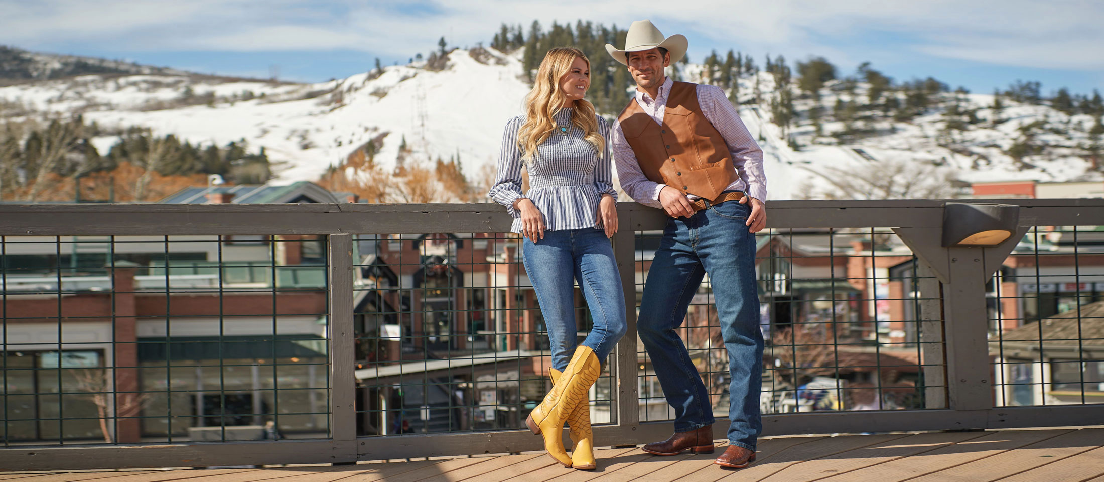 A man and woman standing on a patio wearing cowboy boots. The woman is wearing a blouse shirt and jeans while the man is wearing a shirt, vest, jeans, and cowboy hat.